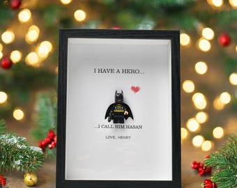 May include: A framed artwork featuring a Lego Batman figure with the text "I HAVE A HERO... I CALL HIM HASAN LOVE, HENRY" and a red heart. The frame is black, and the background is white.