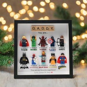 Pode incluir: Uma obra de arte emoldurada com figuras de super-heróis Lego, incluindo Thor, Hulk, Deadpool, Capitão América, Gavião Arqueiro, Batman, Homem-Aranha, Homem de Ferro, Wolverine e Superman. O texto diz "DADDY, You are..." e "You are our favorite superhero! Love, James & Amy."