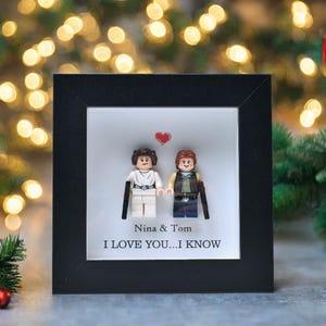 May include: A framed artwork featuring Lego figures of a couple, with a red heart above them. The text reads "Nina & Tom" and "I LOVE YOU...I KNOW". The frame is black, and the background is a blurred display of Christmas lights.