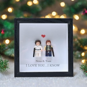 Op de afbeelding: Een zwart omlijst kunstwerk met Lego-figuren van een stel en een rood hart. De witte achtergrond heeft de tekst "Nina & Tom I LOVE YOU...I KNOW". Het kunstwerk staat tegen een vage achtergrond van kerstverlichting.