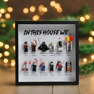 Può includere: Opera d'arte incorniciata con personaggi Lego e la scritta "IN THIS HOUSE WE...". I personaggi rappresentano diversi temi, con frasi come "Dream Big", "Love Friday" e "Eat Good Food". La cornice è nera.