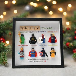 以下が含まれることがあります： レゴのスーパーヒーローのフィギュアが飾られた額縁。フレームには「DADDY, YOU ARE...」というテキストと、「STRONGER THAN」や「SMARTER THAN」などのフレーズが含まれています。下部のテキストは「You are our favorite superhero! Love, James & Amy.」と書かれています。