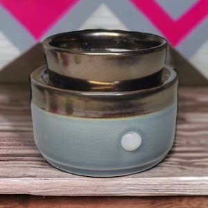 May include: A two-piece ceramic wax warmer. The top piece is a dark bronze color, and the bottom piece is light blue with a crackle finish. A white button is on the front of the base.
