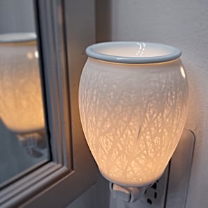 May include: A white ceramic wax warmer with a delicate floral pattern. The warmer is plugged in and lit, casting a warm glow.