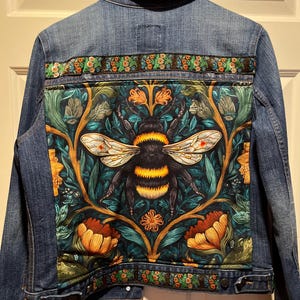 Puede incluir: Chaqueta vaquera con un gran diseño de abeja y flores en la espalda. La abeja es negra y amarilla con alas blancas, sobre un fondo floral verde oscuro y naranja. La chaqueta tiene un ribete floral.
