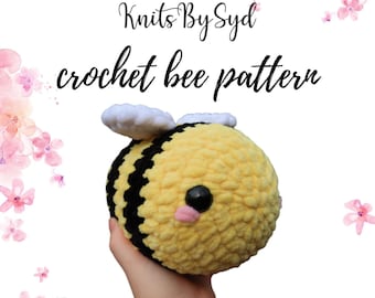 Puede incluir: Un patrón de crochet de abeja con un cuerpo rayado amarillo y negro, alas blancas y mejillas rosadas. La abeja está siendo sostenida en una mano. La imagen tiene un fondo de acuarela con flores rosas.