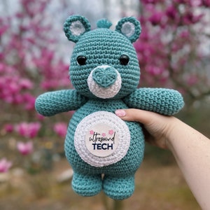 Puede incluir: Un oso de peluche de crochet azul turquesa con un círculo blanco en su vientre que dice "ultrasound TECH" en letras rosa y azul. El oso sostiene una flor rosa.