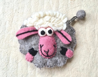 May include: A handmade felt sheep-shaped coin purse. The sheep has a gray body, pink face and ears, and white wool. The coin purse has a zipper and a small gray ball attached. The sheep's face has large, round eyes and a smiling mouth.