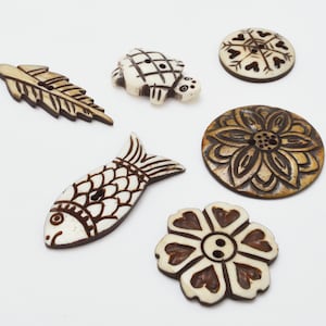 Può includere: Assortimento di sei bottoni in legno con vari disegni, tra cui un pesce, una tartaruga, un fiore, un fiocco di neve e due foglie. I bottoni sono tutti intagliati nel legno e hanno un colore marrone naturale.