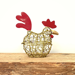May include: A gold wire chicken figurine with red accents. The chicken is standing on a wooden surface.