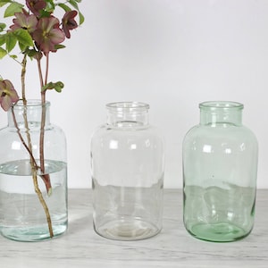 Könnte beinhalten: Drei Glasbehälter in verschiedenen Größen und Farben. Der Behälter links ist klar und enthält einen Zweig mit rosa Blüten. Der mittlere Behälter ist klar und der Behälter rechts ist hellgrün.