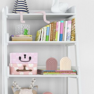 Op de afbeelding: Een witte boekenkast met vier planken, met een verscheidenheid aan boeken, decoratieve items en kaarsen. De bovenste plank heeft een wit zwanenbeeldje en een stapel kleurrijke boeken. De tweede plank heeft een roze en witte opbergdoos, een roze en witte decoratieve doos en een roze en witte kaars. De derde plank heeft een roze en witte kaars en een stapel boeken. De onderste plank heeft een roze en wit konijnenbeeldje en een roze en witte bal.
