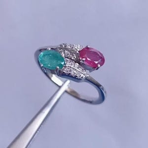 Könnte beinhalten: Ein silberner Ring mit einem grünen Smaragd und einem roten Rubin. Der Ring ist mit kleinen Diamanten besetzt.