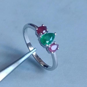 Pode incluir: Um anel de prata com três pedras preciosas em forma de pêra. A pedra central é verde, e as duas pedras de cada lado são vermelhas.