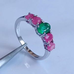 Peut inclure: Une bague en argent ornée d'une rangée de pierres précieuses ovales. La bague alterne des pierres roses vives et vert émeraude. La bague est tenue par une pince.