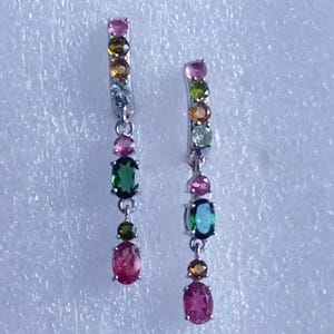 May include: Dangling earrings with a silver-tone metal setting. Each earring features a vertical arrangement of colorful gemstones, including pink, green, yellow, and blue, in various shapes. The gemstones are oval and round.