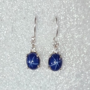 May include: A pair of silver drop earrings featuring oval, deep blue gemstones with a star-like pattern. Each earring has a small cluster of clear stones above the blue gem, and a simple silver hook.