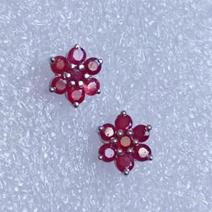 Peut inclure: Deux boucles d'oreilles à clous en forme de fleur. Chaque boucle d'oreille présente un groupe de petites pierres précieuses rondes, rouge foncé, serties dans un métal argenté. Les pierres sont disposées en forme de fleur.