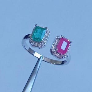 以下が含まれることがあります： エメラルドグリーンと鮮やかなピンクの長方形の宝石が2つ付いたシルバーリング。それぞれの石は、小さな透明な輝く石のハローで囲まれています。リングは、明るい青色の背景に対してピンセットで保持されています。