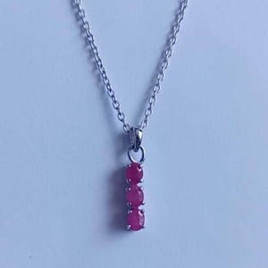 May include: A silver necklace with a pendant featuring three oval, bright pink gemstones. The gemstones are vertically aligned and suspended from a delicate chain. The background is a plain, light color.