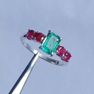 May include: A silver ring featuring a rectangular emerald center stone flanked by three round rubies on each side. The ring is held by a pair of silver tweezers against a light blue background.