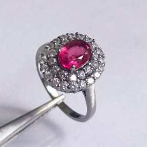 May include: A silver ring featuring a large oval, vibrant red gemstone surrounded by a halo of smaller, clear gemstones. The ring is held by a pair of silver tweezers against a white background. The ring's design is classic and elegant.