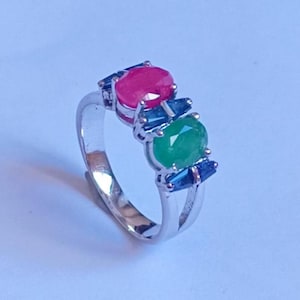 Puede incluir: Un anillo de plata con un diseño llamativo. Presenta un gran rubí ovalado y una esmeralda verde, acentuados por piedras preciosas azules. El anillo tiene una banda ajustable.