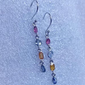 May include: A pair of drop earrings with a sequence of coloured gemstones. Each earring has a hook and a chain of pink, blue, orange, and blue teardrop-shaped stones. The earrings are set against a white background.