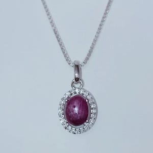 May include: A silver necklace featuring a pendant with a deep red, oval-shaped gemstone, surrounded by a halo of small, clear gemstones. The pendant hangs from a delicate silver chain, creating an elegant and eye-catching piece of jewelry.