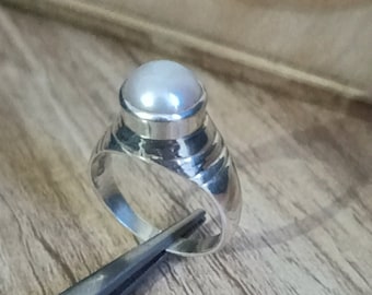 May include: A silver ring featuring a large, round, white pearl set in a bezel. The ring band has a textured design with horizontal grooves. The ring is displayed on a wooden surface.
