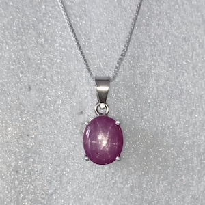 May include: A silver necklace with a pendant featuring a large, oval, purplish-red gemstone. The gemstone has a star-like effect. The pendant is held by four prongs and hangs from a delicate silver chain. The background is a light, textured surface.