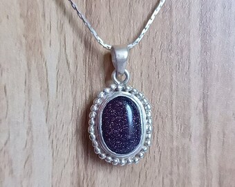 May include: A silver necklace with a dark purple oval pendant. The pendant has a sparkly, textured surface and is surrounded by a silver beaded border.