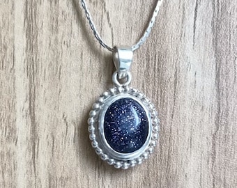 May include: A silver chain necklace with a blue oval pendant. The pendant has a textured surface and is surrounded by a silver beaded border.