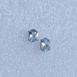 May include: A pair of oval-shaped light blue gemstone stud earrings. Each stone is faceted and secured in a four-prong metal setting, showcasing a clear, translucent appearance with subtle blue tones and reflective surfaces throughout the cut.