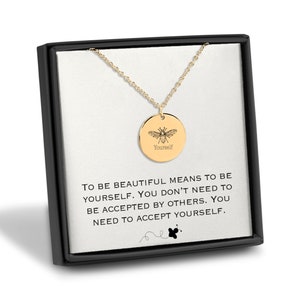 Peut inclure: Un collier en or avec un pendentif rond représentant une abeille et le mot "Yourself" inscrit dessus. Le collier est dans une boîte noire avec un fond blanc et le texte "Être belle signifie être soi-même. Vous n'avez pas besoin d'être accepté par les autres. Vous devez vous accepter vous-même."