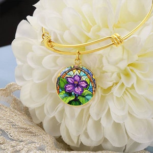May include: A gold expandable bangle bracelet with a round charm featuring a stained glass style violet flower with purple, blue, green, and yellow colors.