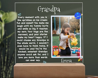 Peut inclure: Un cadre photo lumineux avec une photo d'une jeune fille jouant avec des jouets dans un parc. Le cadre a une base en bois et un devant en acrylique transparent. Le texte sur le cadre dit "Grandpa" et "Every moment with you is like sprinkles on ice cream: fun and sweet! You make me laugh until my tummy hurts and smile so big it reaches my ears. Your hugs are the warmest, and your stories make my heart happy. If I could choose any Grandpa in the whole world, I wouldn't even have to think twice, it would be you! You're the kindest, sweetest Grandpa anyone could ask for, and I love you more than words can ever say." Le texte comprend également un symbole de cœur et un symbole de fusée. Le nom de la fille est "Emma".