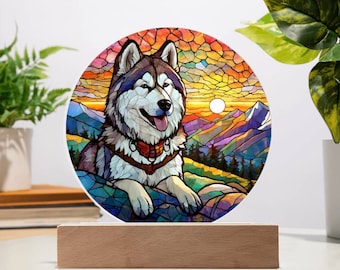 May include: A stained glass style illustration of a husky dog sitting in a mountain landscape with a sunset in the background. The dog is looking at the viewer with its tongue sticking out. The image is circular and has a wooden base.