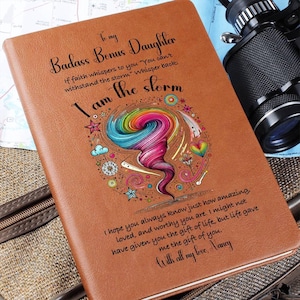 Op de afbeelding: Een bruin lederen notitieboek met een kleurrijk tornado-ontwerp en de tekst "To my Badass Bonus Daughter If faith whispers to you "You can't withstand the storm" Whisper back: I am the storm" en "I hope you always know just how amazing, loved, and worthy you are. I might not have given you the gift of life, but life gave me the gift of you. With all my love, Nancy."