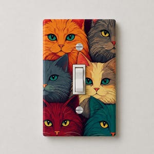 May include: A rectangular light switch cover plate featuring a colorful illustration of six cat faces in orange, gray, blue, red, and teal. The plate has a central vertical toggle switch and two silver screws at the top and bottom for mounting.