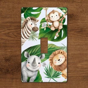 May include: A decorative light switch cover featuring a jungle theme. The design includes a zebra, monkey, rhino, and lion, surrounded by green tropical leaves. The background is white.
