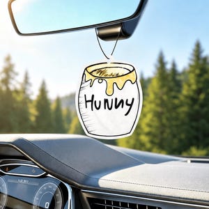 May include: A white car air freshener shaped like a honey pot with yellow honey dripping from the rim. The word "HUNNY" is printed in black, handwritten-style letters on the front of the pot. It hangs from a thin black string attached to a rearview mirror.