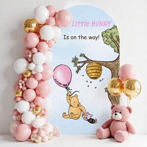 May include: A baby shower decoration featuring a pink teddy bear, a balloon arch with pink, white, and gold balloons, and a backdrop with Winnie the Pooh illustrations and the text "Little Hunny Is on the way!"