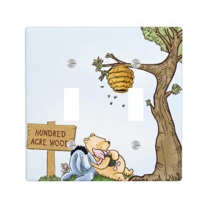 May include: A double light switch cover featuring a cartoon illustration of a yellow bear and a blue donkey sitting under a tree with a beehive. A wooden sign in the foreground reads HUNDRED ACRE WOOD. The background is a light blue sky.