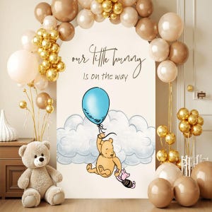 May include: A baby shower decoration featuring a Winnie the Pooh theme. The scene includes a large sign with the text "our little bunny is on the way", a cartoon of Winnie the Pooh holding a blue balloon, and a plush teddy bear. Balloons in gold, white, and brown complete the decor.