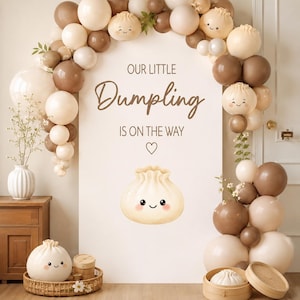 May include: A baby shower decoration featuring a white backdrop with the text "OUR LITTLE Dumpling IS ON THE WAY." The backdrop is decorated with a balloon arch in shades of brown and white. A dumpling illustration is in the center.