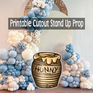 May include: A printable cutout stand-up prop featuring a cartoon honey pot with the word "HUNNY" and a balloon arch in shades of blue and white. The prop is designed for parties and events.