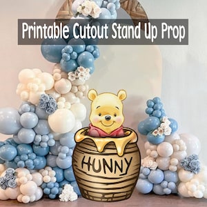 May include: A printable cutout stand-up prop featuring Winnie the Pooh in a honey pot with the word "HUNNY". The background includes a balloon arch in shades of blue and white, with floral accents. The text "Printable Cutout Stand Up Prop" is at the top.