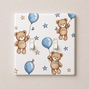 May include: A white double light switch wall plate featuring a repeating pattern of brown teddy bears holding blue balloons, interspersed with small blue and tan stars. The plate includes two white toggle switches and four silver mounting screws.