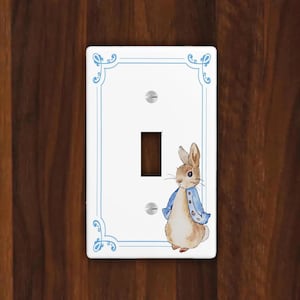 May include: White light switch cover with a blue border and a watercolor illustration of a rabbit wearing a blue jacket. The switch plate has a single toggle switch and two screw holes.